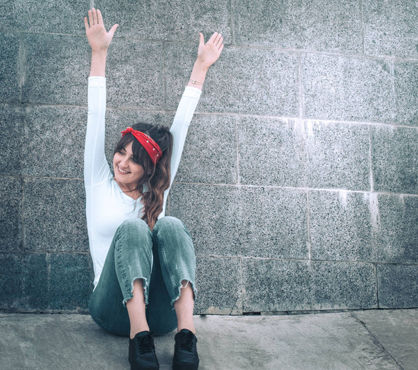 Красивая девушка в джинсах и белой футболке с красной повязкой на голове, на фоне стены, концепция городской одежды и молодежного стиля
 - Фото, изображение