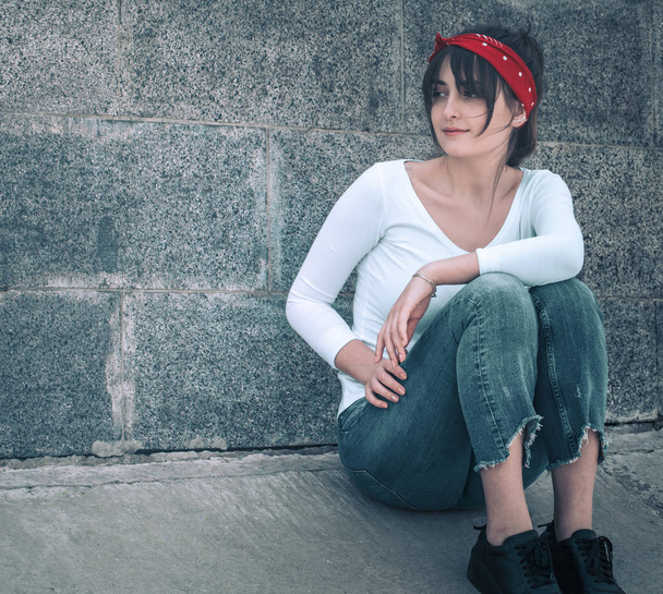 Красивая девушка в джинсах и белой футболке с красной повязкой на голове, на фоне стены, концепция городской одежды и молодежного стиля
 - Фото, изображение
