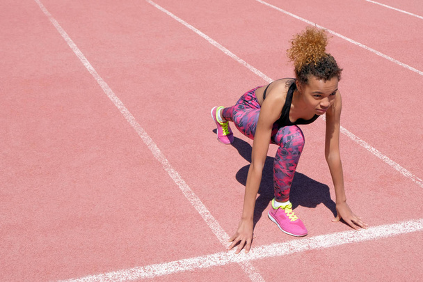 Nuori kaunis afroamerikkalainen tyttö urheilullisessa mustassa t-paidassa ja vaaleanpunaisissa lenkkareissa valmistautuu kisaamaan juoksumaton alkukaistalla. Elämäntapa. Kopioi tila
. - Valokuva, kuva