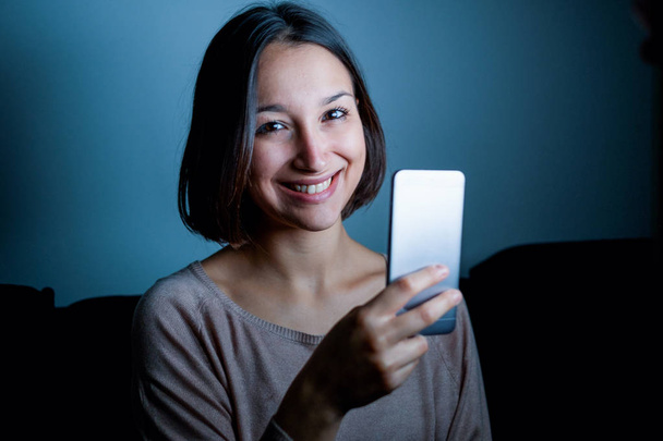 Giovane donna che utilizza il cellulare nel buio di notte guardando la fotocamera
 - Foto, immagini