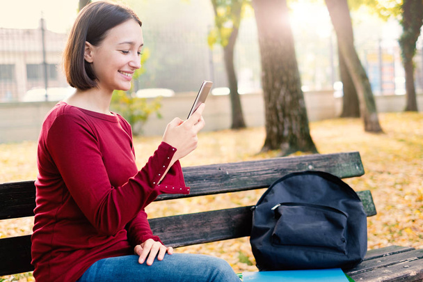 Jonge student meisje te wachten en houden van mobiele telefoon in het stadspark - Foto, afbeelding