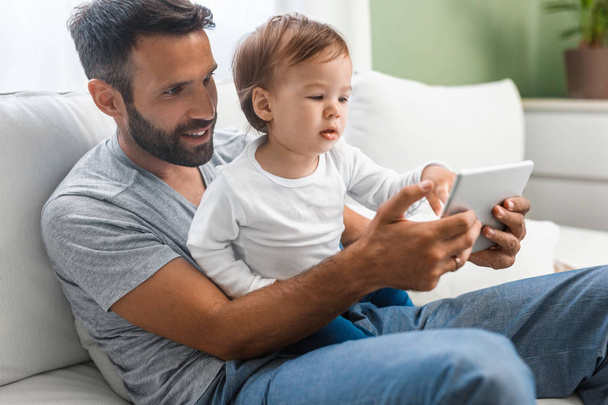 Handsome Caucasian man holding his cute baby son and looking at tablet. - Foto, imagen
