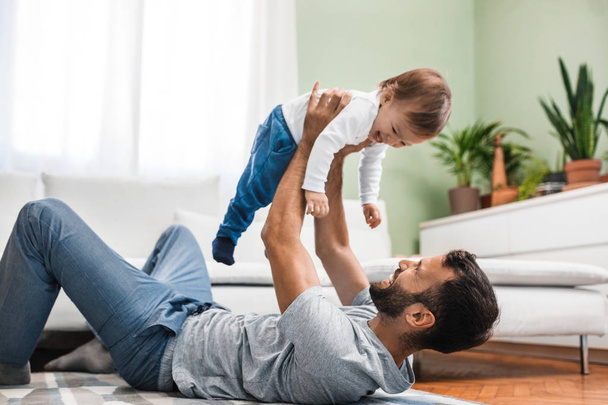 Happy Caucasian man lying on the floor and playing with his baby son. - Φωτογραφία, εικόνα