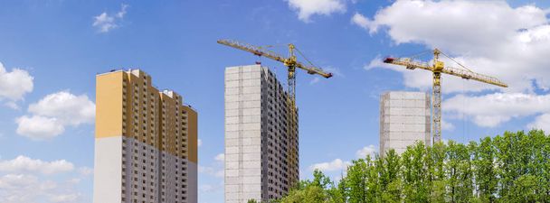 Panorama de la construcción de las casas residenciales de varios pisos a partir de paneles de hormigón prefabricados con grúas torre de construcción y árboles en primer plano contra el cielo
 - Foto, imagen