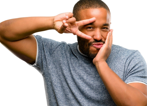 Hombre afroamericano con barba mirando a la cámara a través de los dedos en gesto de victoria guiñando el ojo y soplando un beso aislado sobre fondo blanco
 - Foto, imagen