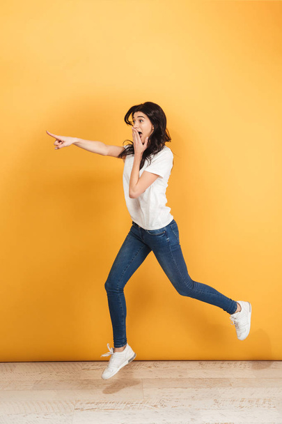 Image of young shocked woman jumping isolated over yellow background. Looking aside pointing. - Foto, immagini