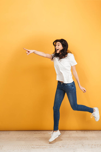 Image of surprised cute young pretty woman jumping isolated over yellow background pointing. - Фото, изображение
