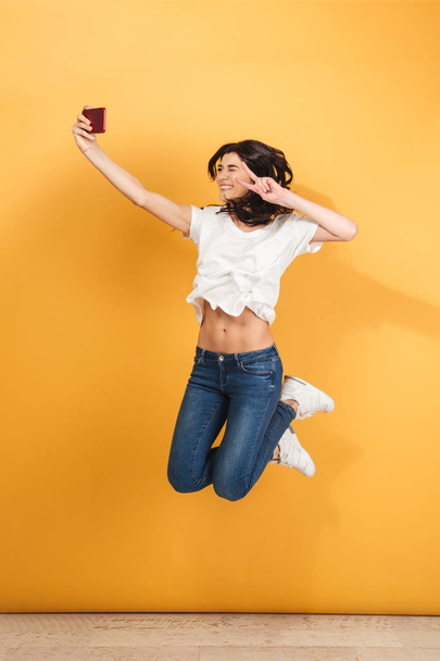 Image of young emotional woman jumping isolated over yellow background make selfie by mobile phone with peace gesture. - 写真・画像