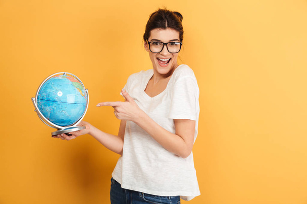Image of young excited happy woman wearing glasses isolated over yellow background holding globe. - Fotoğraf, Görsel