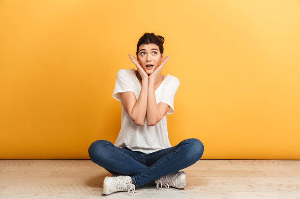 Portrait of a cheerful young woman sitting with legs crossed on a floor and looking away over yellow background - Foto, Imagem