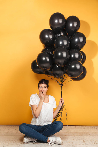 Portrait of an excited young woman holding bunch of black air balloons while sitting with legs crossed on a floor over yellow background - Foto, Bild
