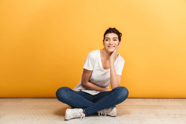 Portrait of a smiling young woman sitting with legs crossed on a floor and looking at camera over yellow background - Fotó, kép