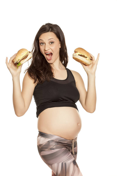 Молода вагітна жадібна жінка тримає двох бургерів на білому тлі
 - Фото, зображення