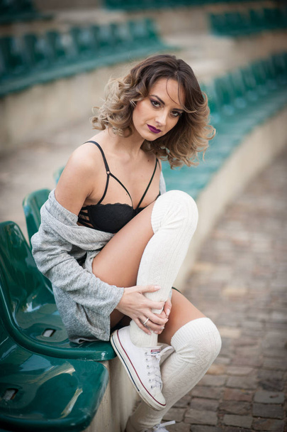 Sensual girl with long legs in the courts of a field .Long legs attractive blonde with curly hair relaxing on the chair in the stands.Fashionable young woman posing sensual with white woolen socks - 写真・画像