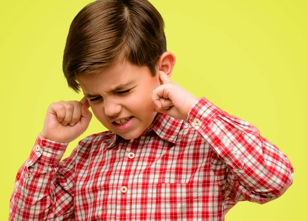 Przystojny przedszkolaków z zielonymi oczami obejmujące uszy, ignorując przykry hałas, podłącza uszy aby uniknąć słyszenie dźwięku. Głośno muzyki jest problem. na żółtym tle - Zdjęcie, obraz