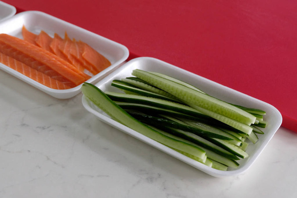 Ingredientes para cocinar sushi y rollos.Pepino fresco y filete de salmón rojo en una taza rectangular blanca junto a una tabla de cortar. Comida japonesa
. - Foto, Imagen