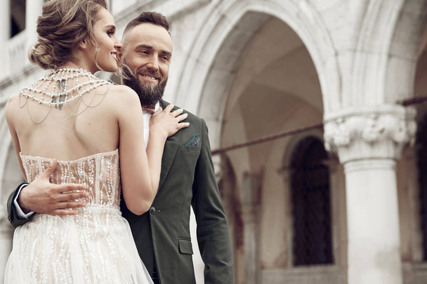 Portrait of tender wedding couple. Bearded man in green suit, beautiful woman with tattoo on back, dressed in luxury elegant wedding dress. St Mark's Square, Venice, Italy - Photo, Image