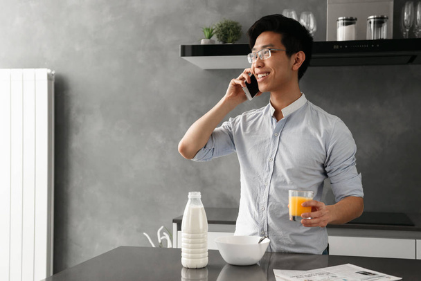 Portrait of a smiling young asian man talking on mobile phone while having breakfast on a kitchen at home - Zdjęcie, obraz