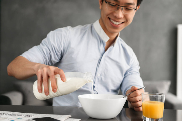 Portrait of a joyful young asian man having breakfast while sitting at the table on a kitchen at home - Foto, imagen