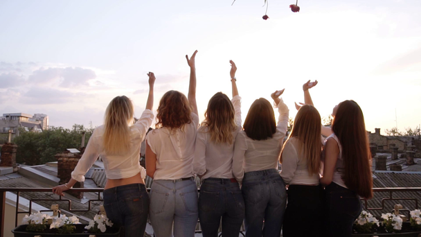 Caucasian girls with beautiful figures, in the same casual clothes standing with their backs to the camera, lean against the balcony railings. throw flowers into the air. Cheerful. Backside. - Footage, Video