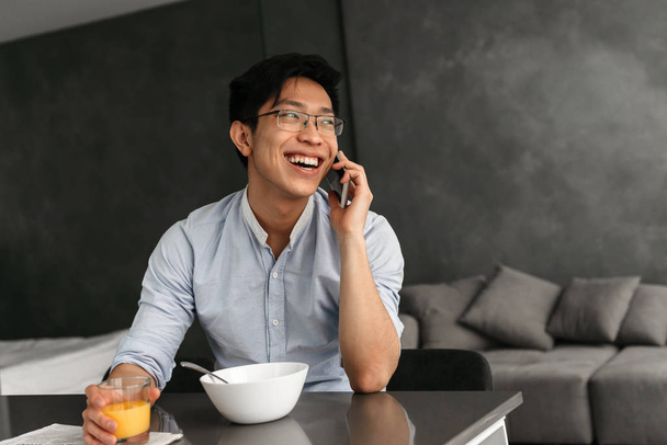 Portrait of a laughing young asian man having breakfast while sitting at the table on a kitchen at home and talking on mobile phone - Fotoğraf, Görsel