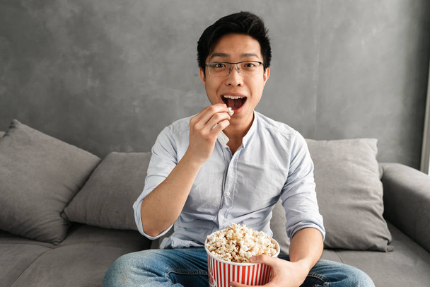Portrait of a happy young asian man eating popcorn while sitting on a couch at home and watching TV - Photo, image