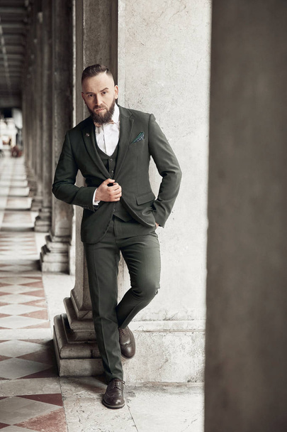 Bearded man in brown green three-piece suit, bow tie and brogue shoes posing near columns in Venice, Italy - Foto, Bild