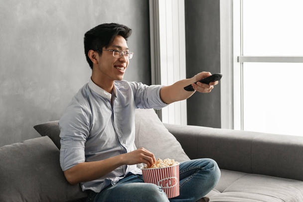 Portrait of a joyful young asian man holding popcorn and TV remote control while sitting on a couch at home - Zdjęcie, obraz
