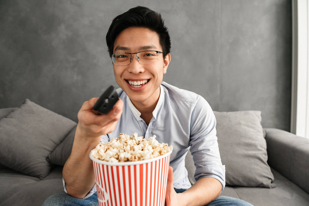 Portrait of a cheerful young asian man holding popcorn and TV remote control while sitting on a couch at home - Foto, immagini