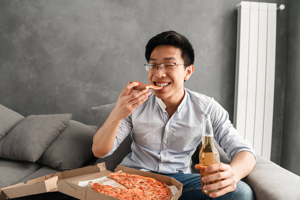 Portrait of a satisfied young asian man eating pizza and drinking beer while sitting on a couch at home and watching TV - Foto, immagini