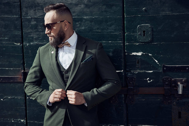 Bearded adult man in dark sunglasses, dressed in brown green three-piece suit, bow tie, posing near green gate in Venice - Photo, Image