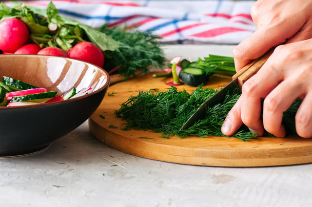 Здоровый салат из свежих овощей в процессе приготовления. Женские руки режут укроп на деревянной доске. Пошаговая концепция приготовления пищи
. - Фото, изображение