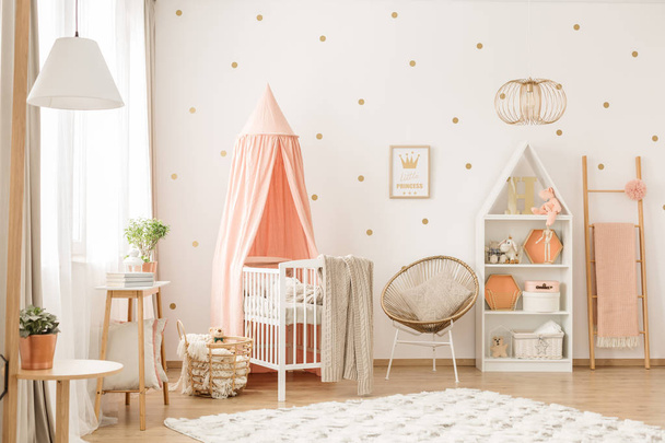 Gold chair with pillow standing next to a white wooden crib with pastel pink canopy in bright baby room interior with dots on the wall - Foto, immagini