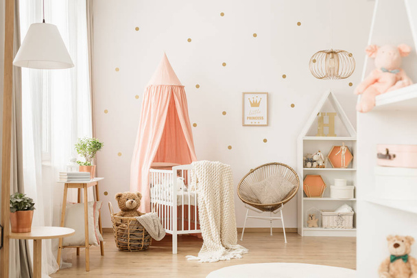 Canopied cradle between gold armchair and basket with teddy bear in pink baby's bedroom interior - 写真・画像