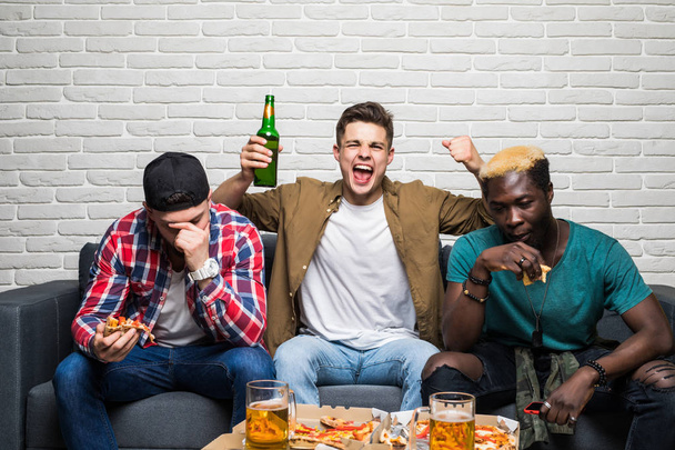 drei attraktive, gut aussehende, emotionale Männer fernsehen, sich Sorgen machen, jubeln, mit dem Zeigefinger zeigen, Pizza, Pils, Alkohol im Wohnzimmer essen - Foto, Bild