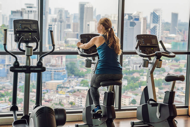 Young woman on a stationary bike in a gym on a big city background. - Фото, изображение