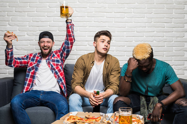 Three mixed race men sitting on sofa drink beer and eat pizza support different team and with different emotion of lose and win at home - Photo, Image