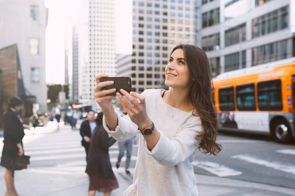 Kaunis nuori nainen bulevardilla kaupunkimaisemassa, keskustassa, auringonlaskun aikaan, pitämällä älypuhelinta ja ottamalla valokuvia
. - Valokuva, kuva