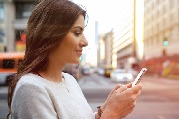Mooie jonge vrouw op de boulevard in stedelijke landschap, centrum, bij zonsondergang, bezit smartphone - Foto, afbeelding