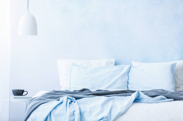 Blue bedsheets and pillows on bed next to a table with a cup under white lamp in simple bedroom interior - 写真・画像