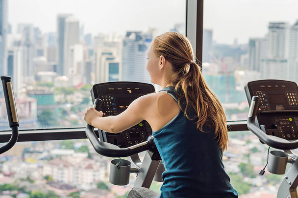 Young woman on a stationary bike in a gym on a big city background. - Foto, Imagen