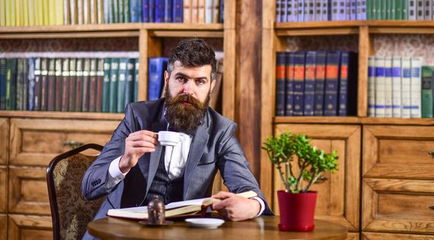 man reading bookBusinessman sits in vintage interior, holds book and cup of tea. Bearded man in luxury suit in his cabinet. Mature man with calm face enjoys reading. Luxury lifestyle, vintage fashion - Fotoğraf, Görsel
