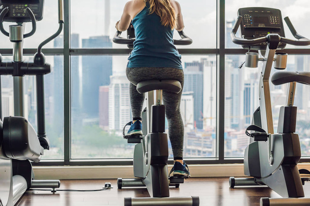 Young woman on a stationary bike in a gym on a big city background. - Foto, Imagem