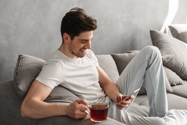 Handsome man in basic t-shirt smiling and holding mobile phone in hands while drinking tea on couch in house - Fotoğraf, Görsel