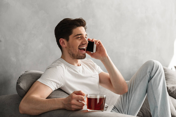 Young and beautiful man in basic t-shirt smiling and speaking on mobile phone while drinking tea on couch in house - Photo, Image