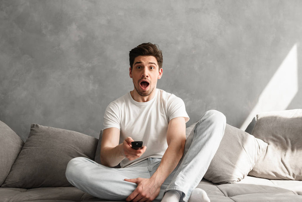 Photo of excited man with open mouth sitting on couch in gray apartment and holding remote control for tv in hand - Photo, Image