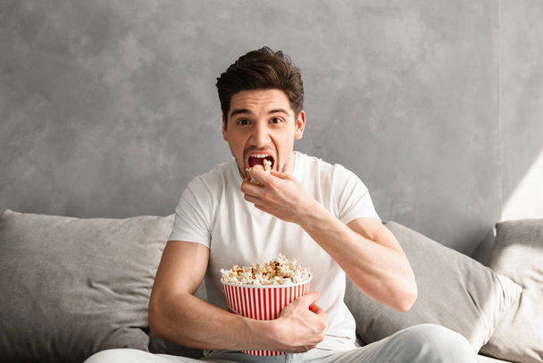 Single man 20s sitting on sofa in gray apartment and eating pop corn while watching tv in morning - 写真・画像