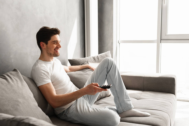 Image in profile of attractive brunette man sitting on sofa at home and watching television with remote control in hand - Valokuva, kuva