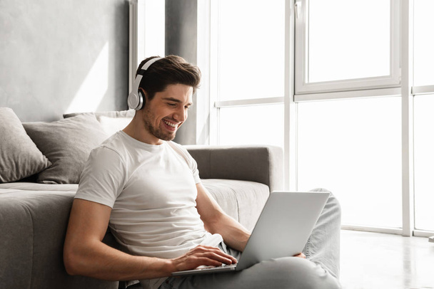 Bearded man 30s in basic clothing sitting on floor at home and listening to music on laptop wearing wireless headphones - Valokuva, kuva