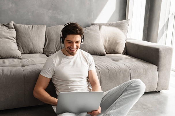 Happy guy with dark hair sitting on floor indoor and listening to music on laptop with wireless headphones - Photo, image